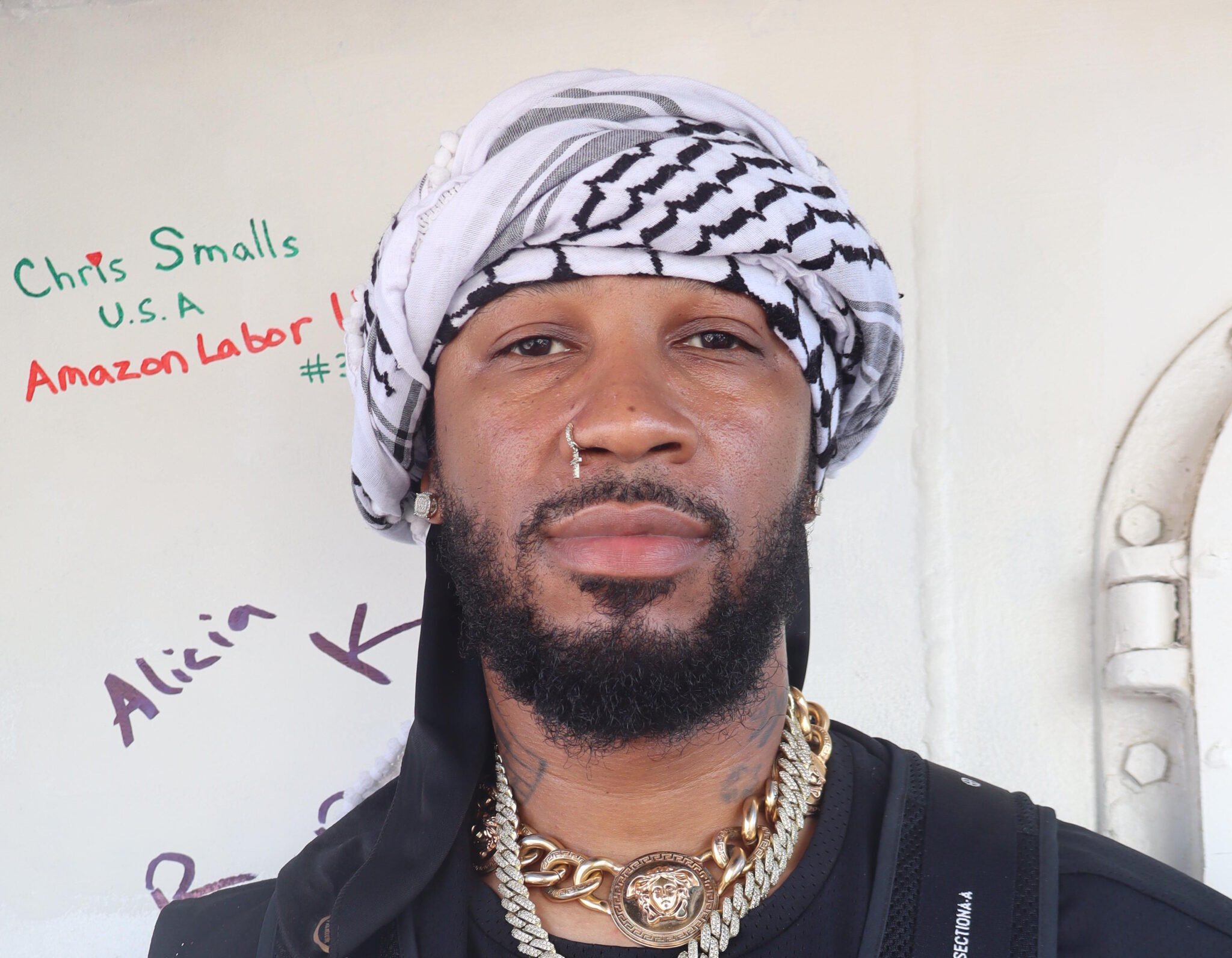 a black person with a scruffy beard and mustache and cross piercing in one nostril, wearing a kuffiyeh head covering and gold necklaces