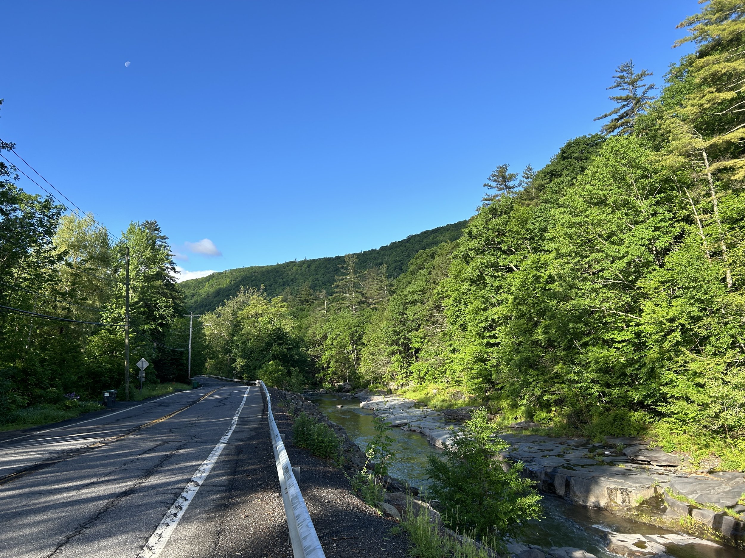 a creek and 2-lane road running parallel to each other with green trees on either side and a blue sky with waning moon