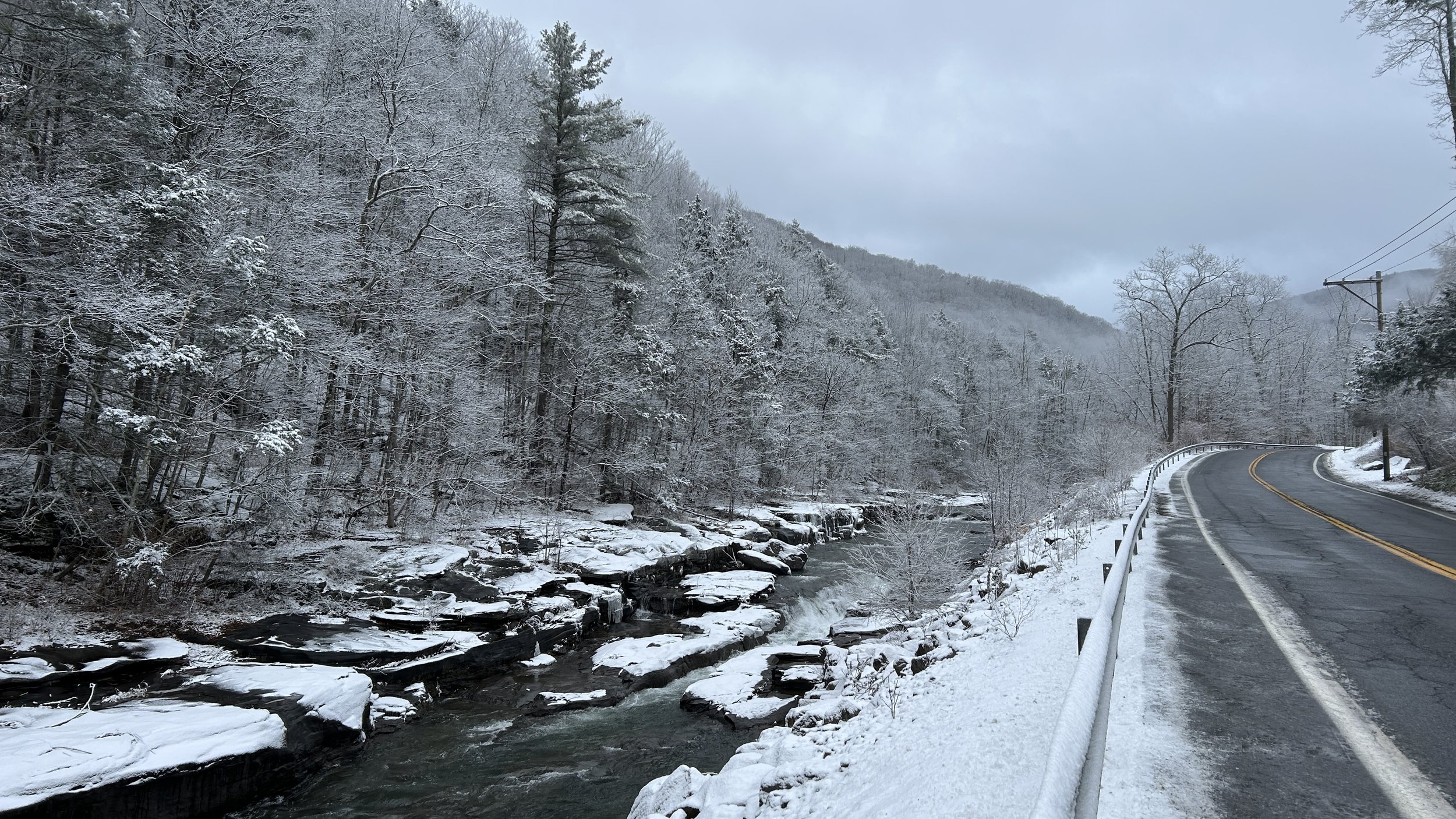 a snow-covered landscape with a creek running parallel to a two-lane road