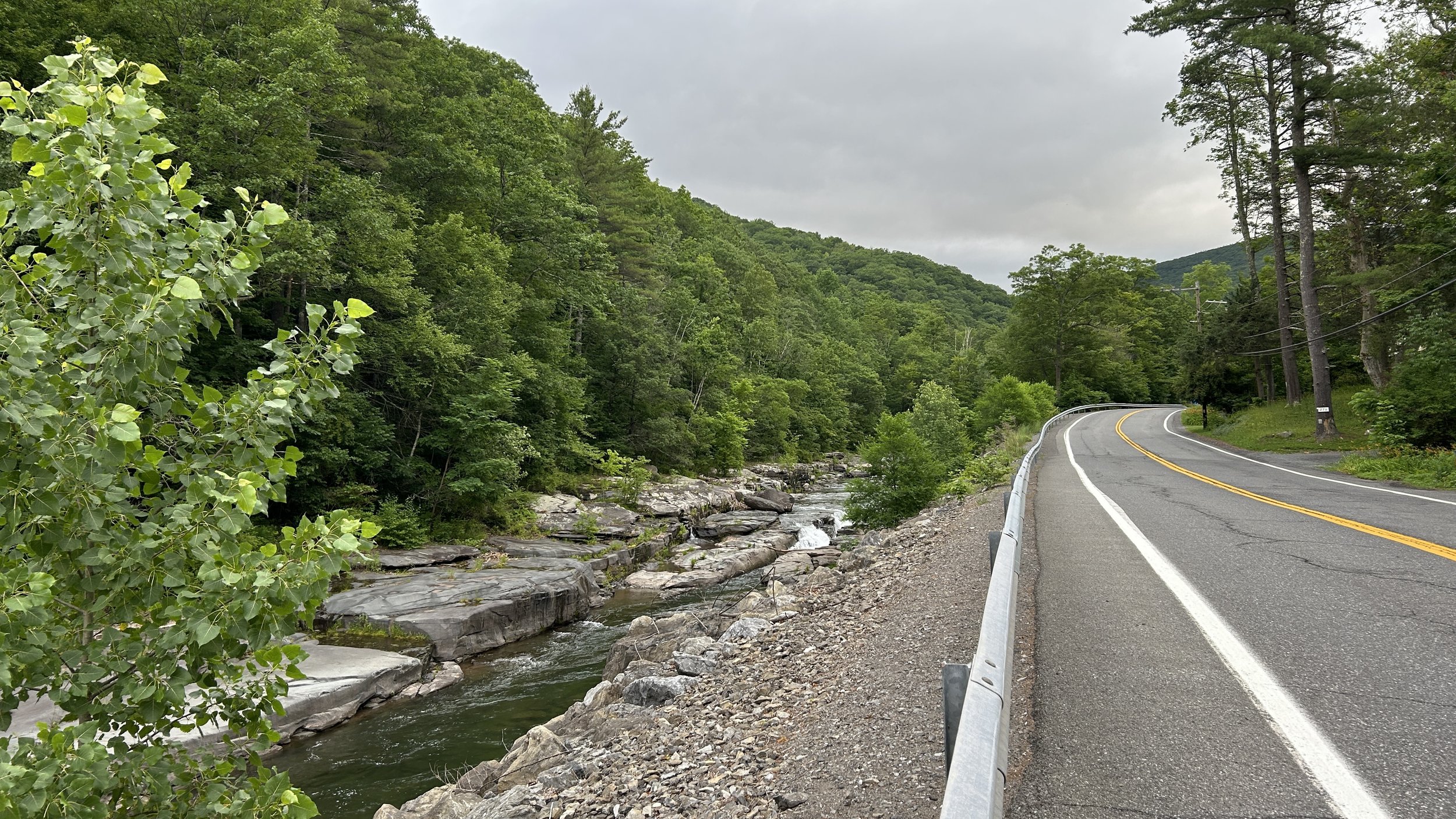 a stream running thru flat grey rocks bordered by green trees on one side and a 2-lane road on the other