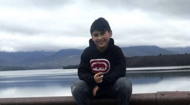 a 9-year-old child with light brown skin and brown hair wearing a hooded sweatshirt sitting on a metal railing in front of a body of water and misty mountains