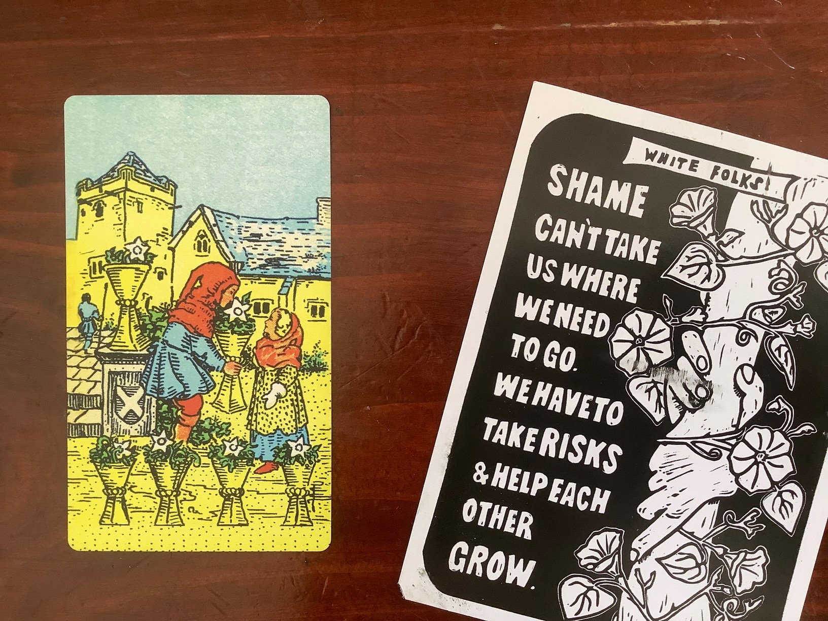 a tarot card and a black and white postcard that says White Folks Shame can't take us where we need to go. We have to take risks and help each other grow, against a dark wood background