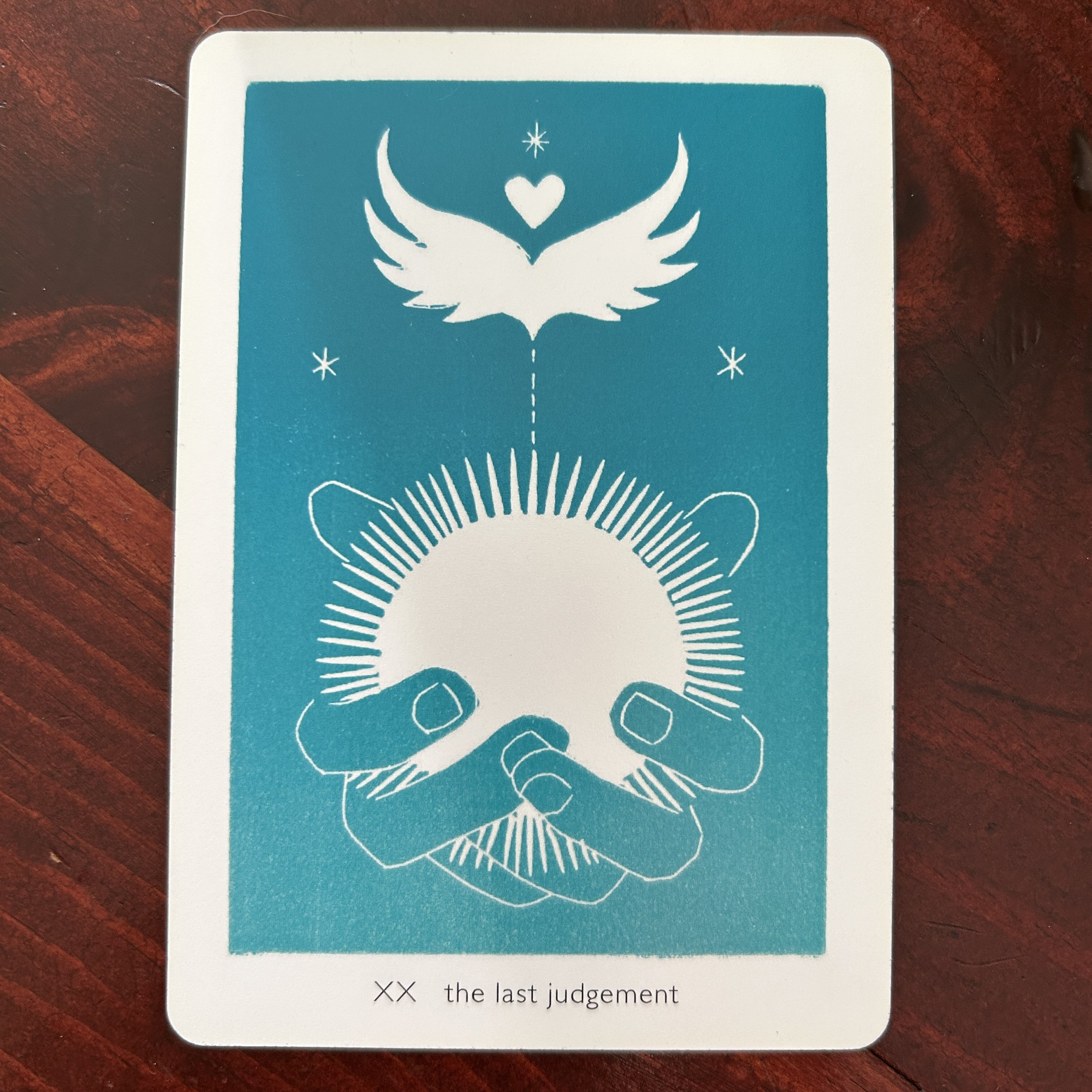 linocut print of teal-blue hands gently holding a white radiating circle above which white wings, heart and stars hover against a teal-blue ground