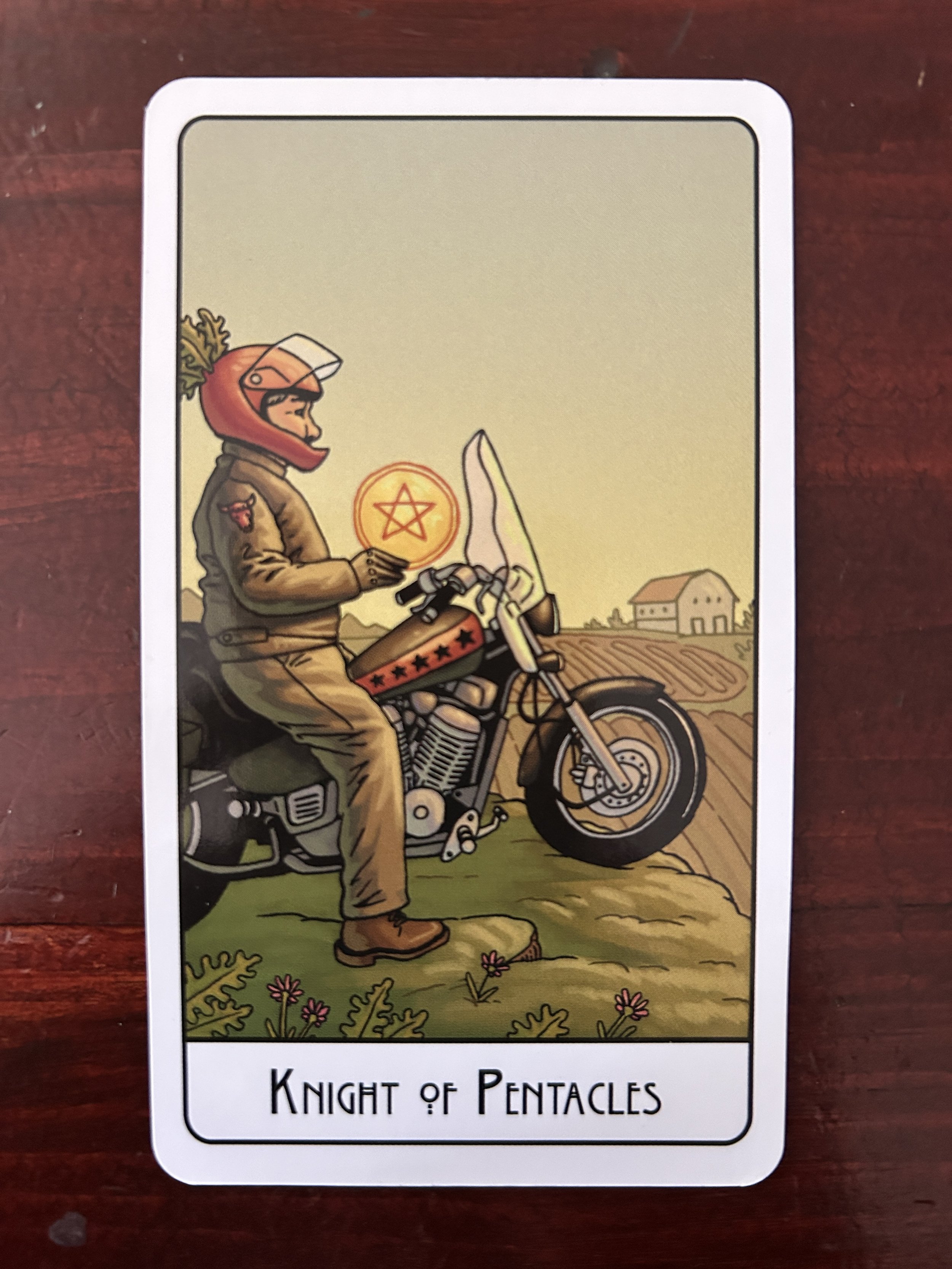 A person in profile wearing a helmet with the visor open sits on a still motorcycle with a red stripe and black stars. a field, barn and big sky are in the background