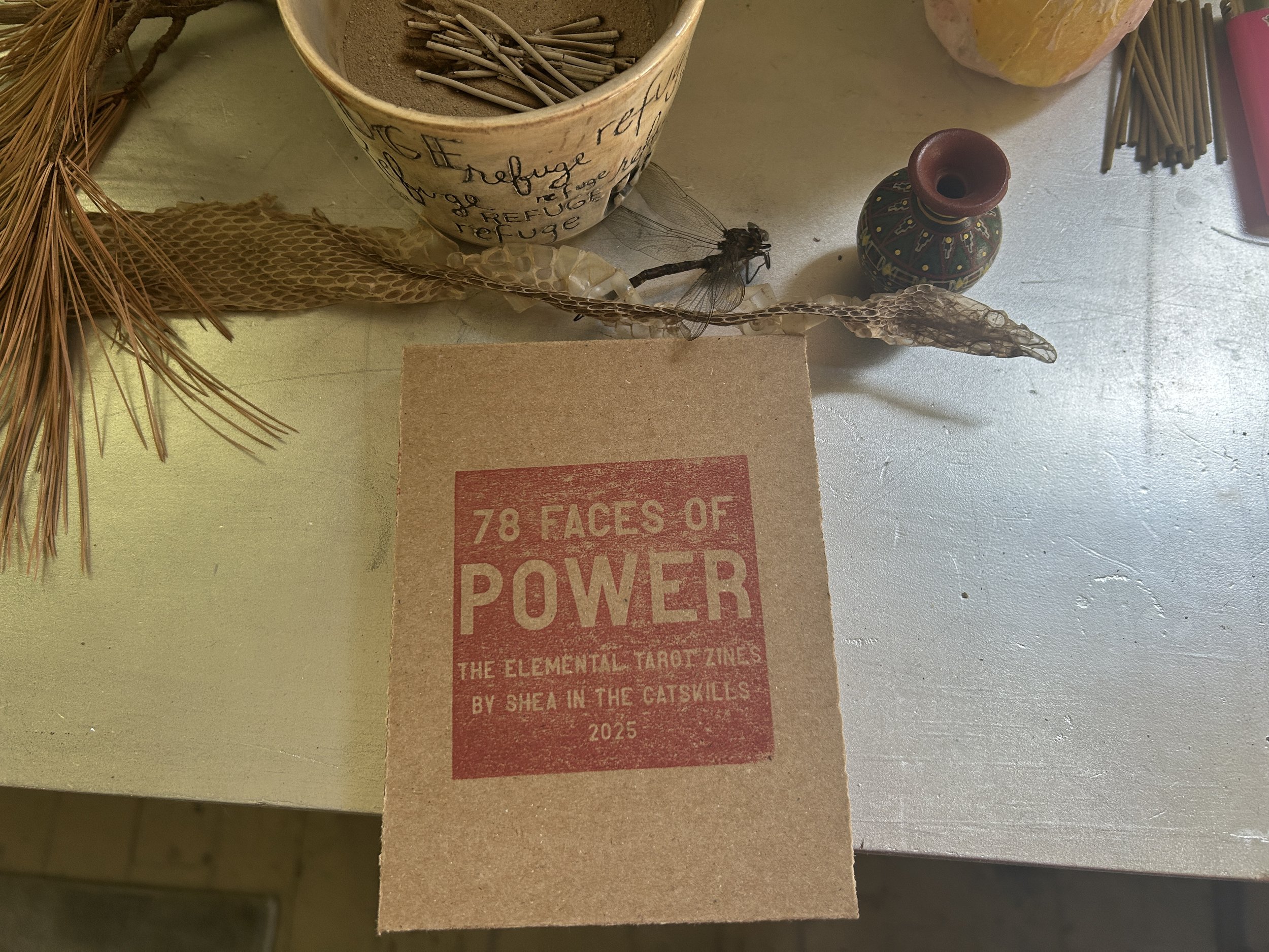 red-stamped cardboard cover of the zine box set on a silver altar with an incense bowl, dragonfly carcass and snake skin 