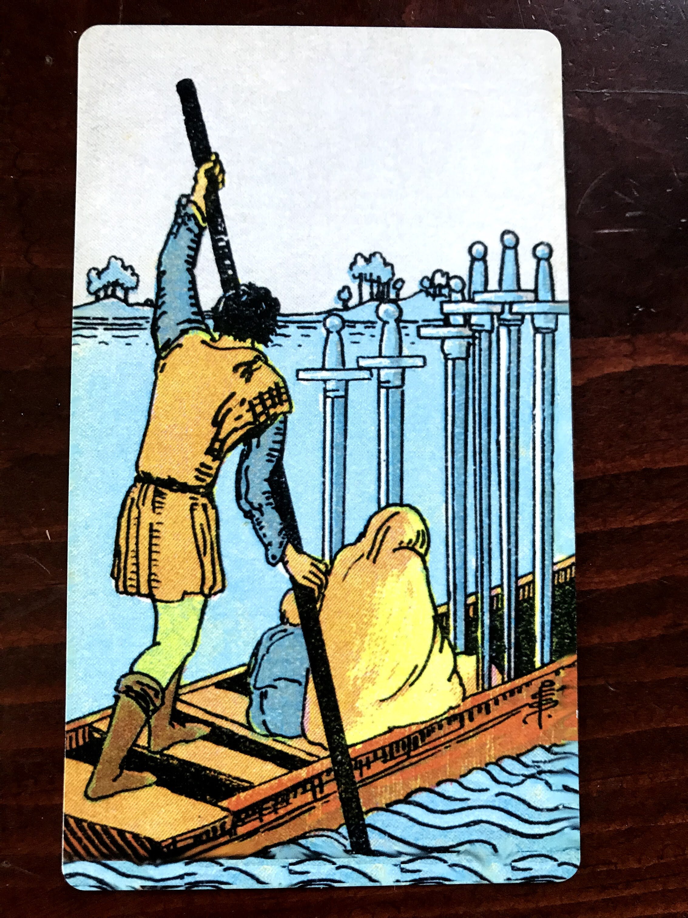 a tarot card depicting 2 figures in a boat with 6 swords being ferried by a standing figure across blue water