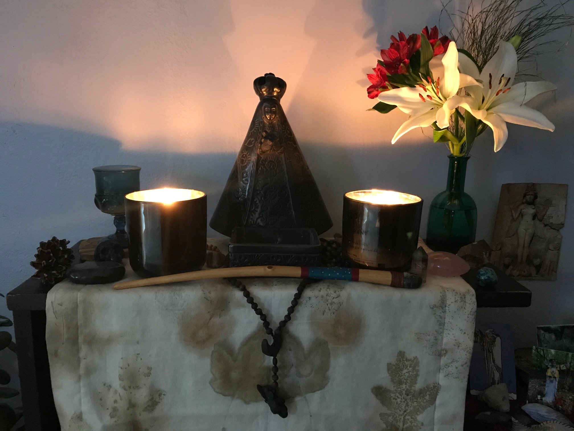 an altar with two lit candles, a wand, a rosary, an ecoprinted altar cloth, a black ceramic statue of Mary and a vase of fresh flowers
