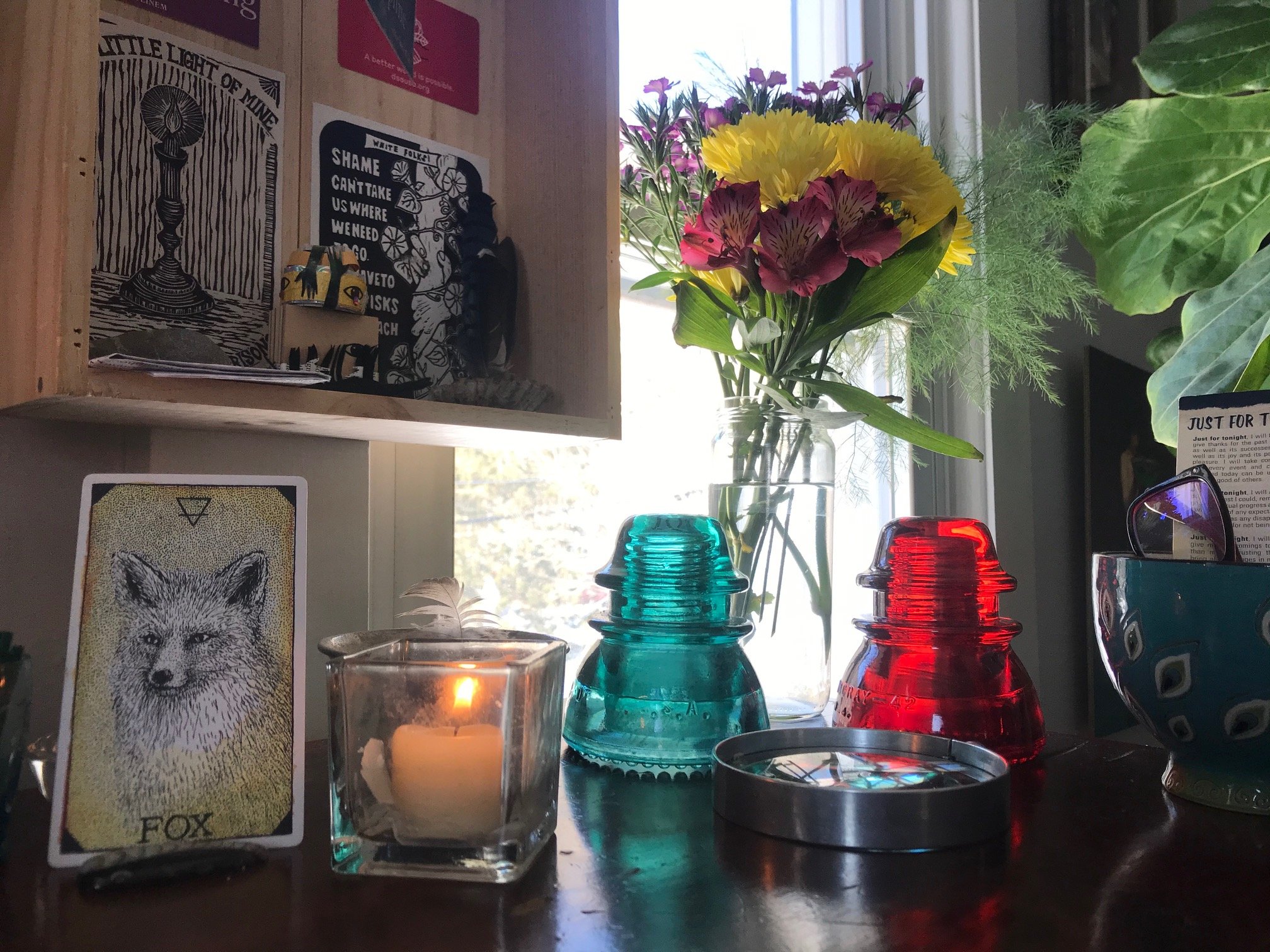 an altar on a desk that includes a small lit candle a fox oracle card, red and blue glass telephone pole insulators, flowers, and a peacock patterned teacup with reading glasses in it