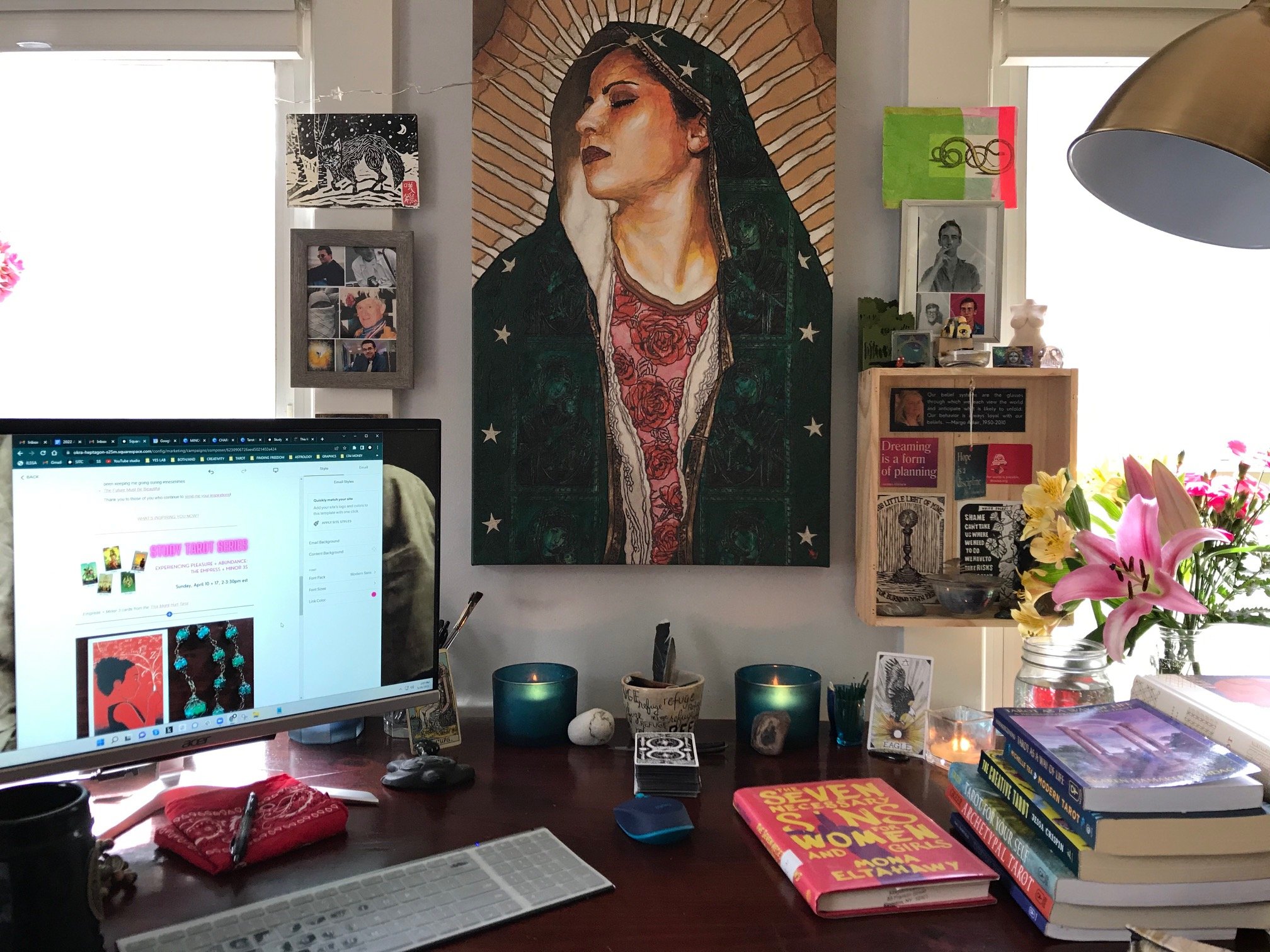 an image of a work altar on a desk that includes a computer, stacks of books, candles, flower artwork and a large print of the Virgin of Guadalupe