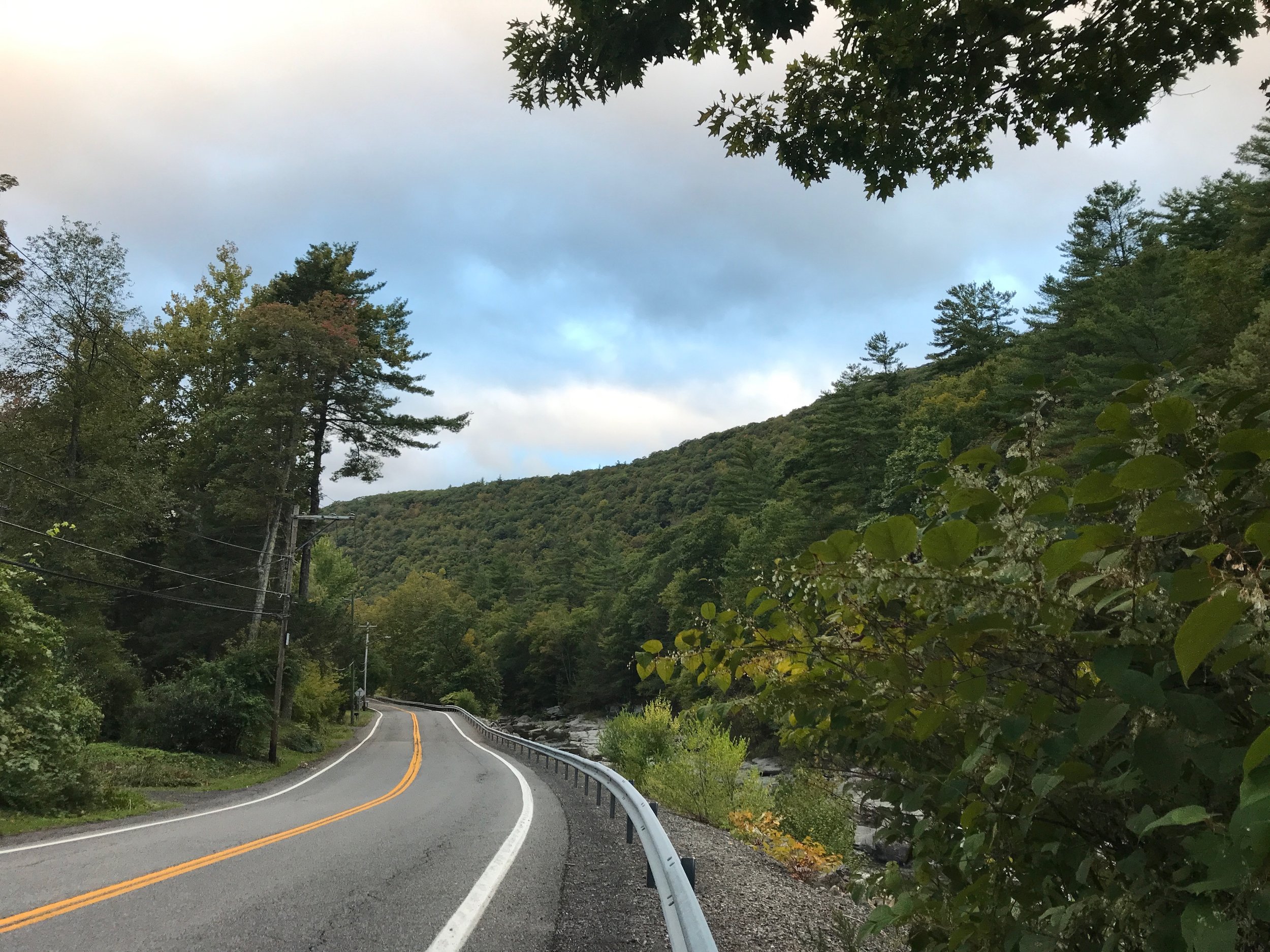 a two-lane road with a double yellow line running along a creek, flanked by lush green trees and a mountain, a cloudy blue sky
