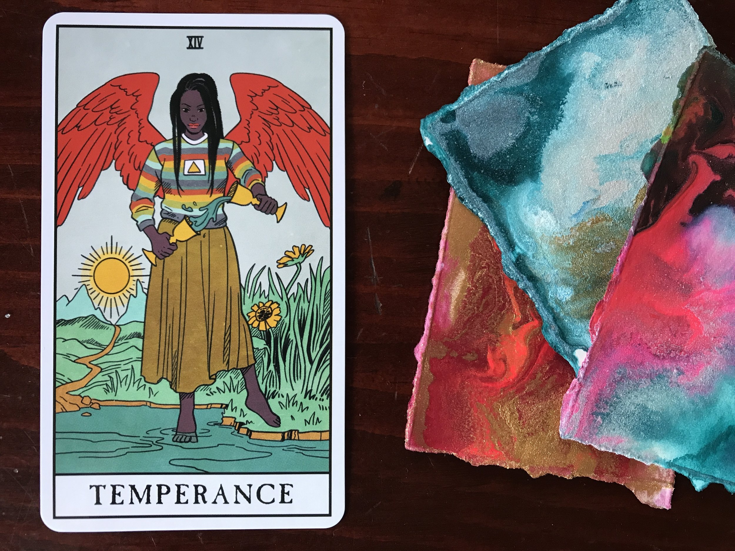 a tarot card and a pile of small brightly colored abstract paintings against a dark wood background