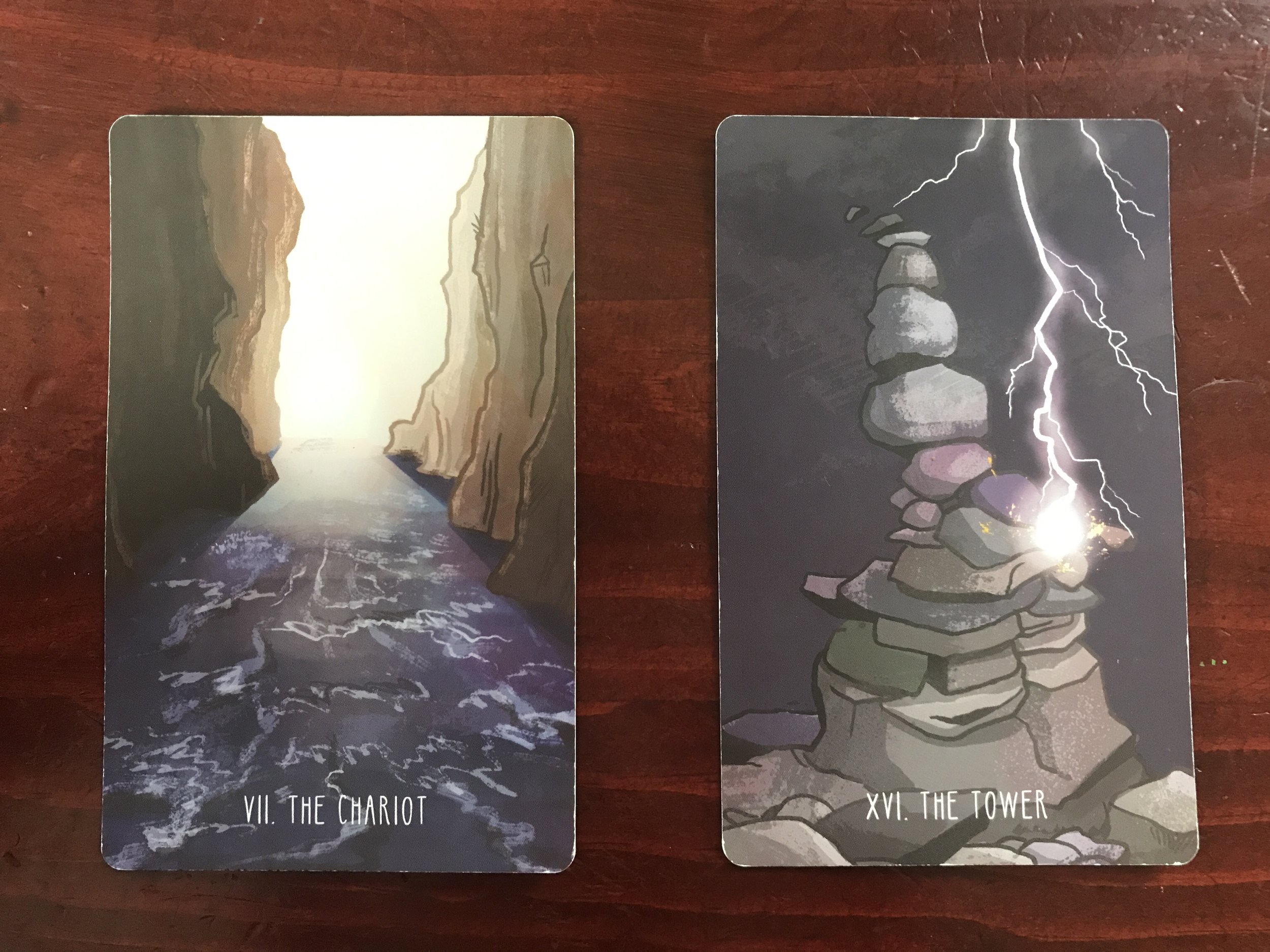 2 tarot cards against a dark wood background, one showing a river running through a canyon, the other showing a bolt of lightning striking a cairn