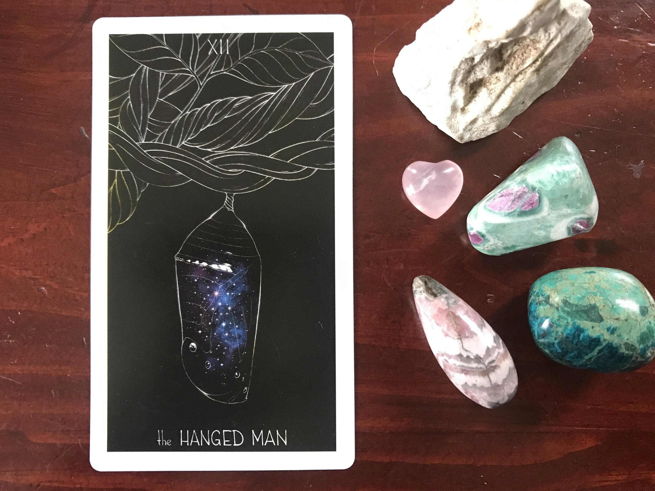 a tarot card showing a blue starry chrysalis against a dark wood background with colored stones