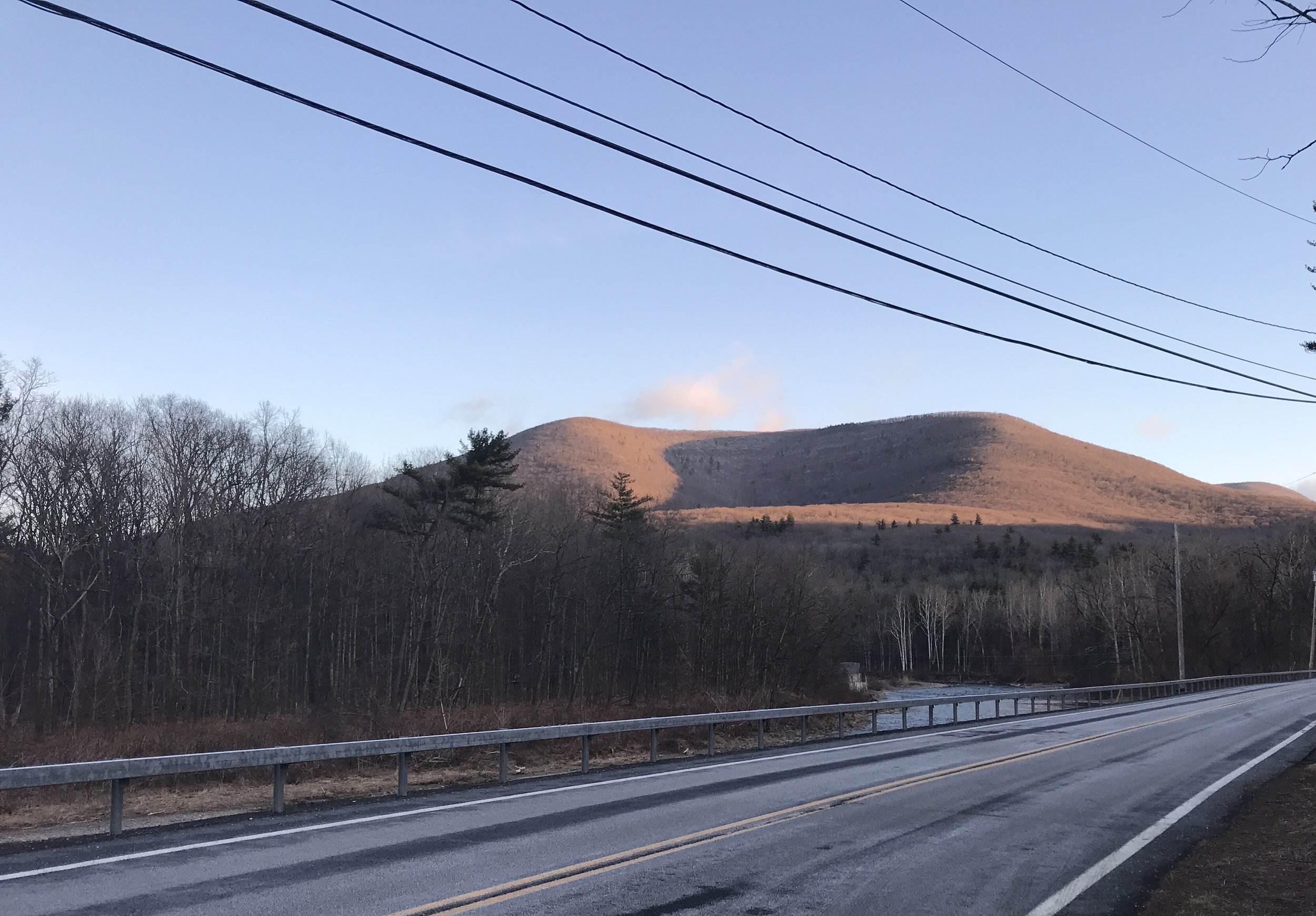 a bare winter landscape of trees, a river and a two-peaked mountain with deep shadow between the peaks and a pale blue sky