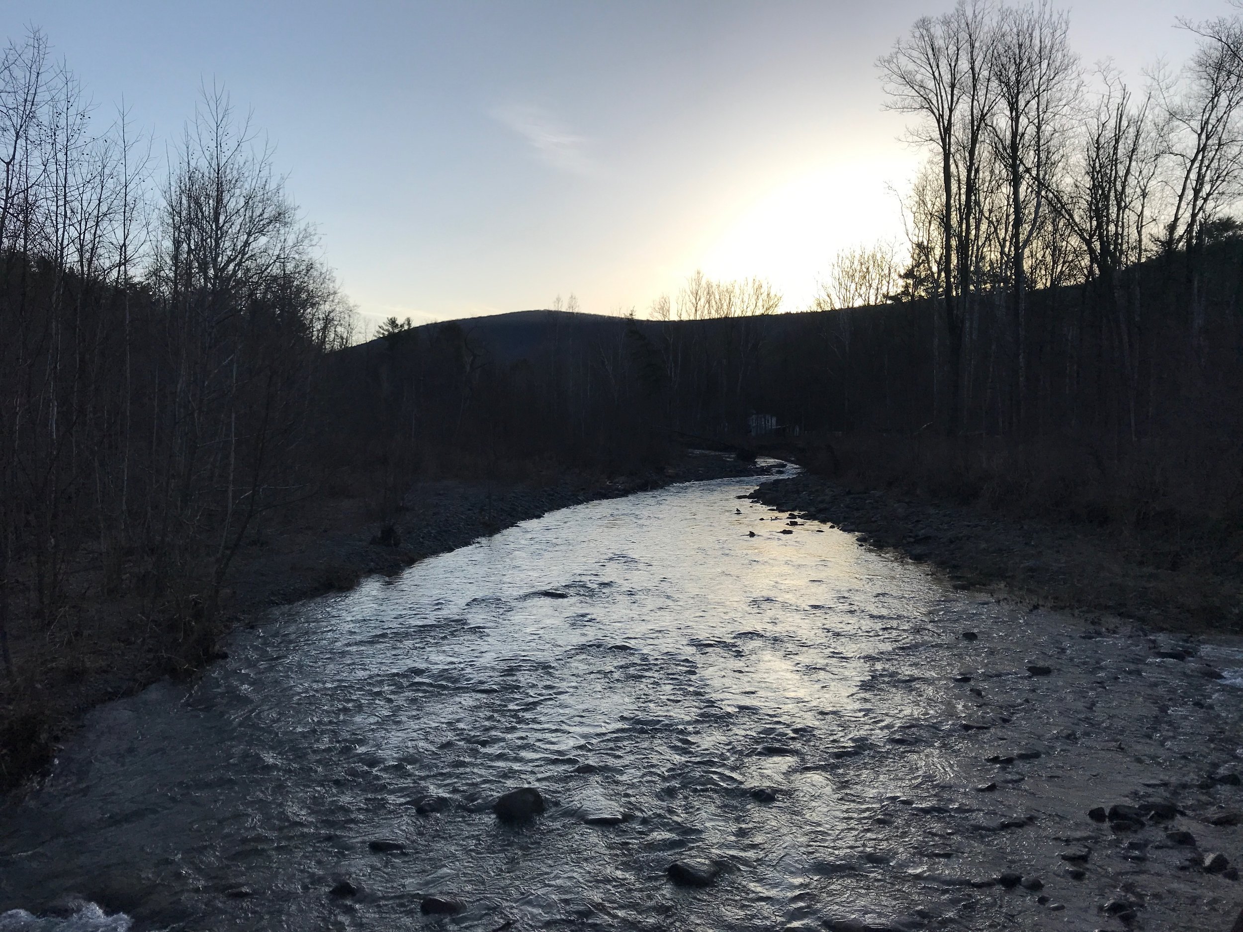 a winding creek, bare trees and the shape of a mountain in the stark contrast light of dawn as the sun rises in a pale blue sky