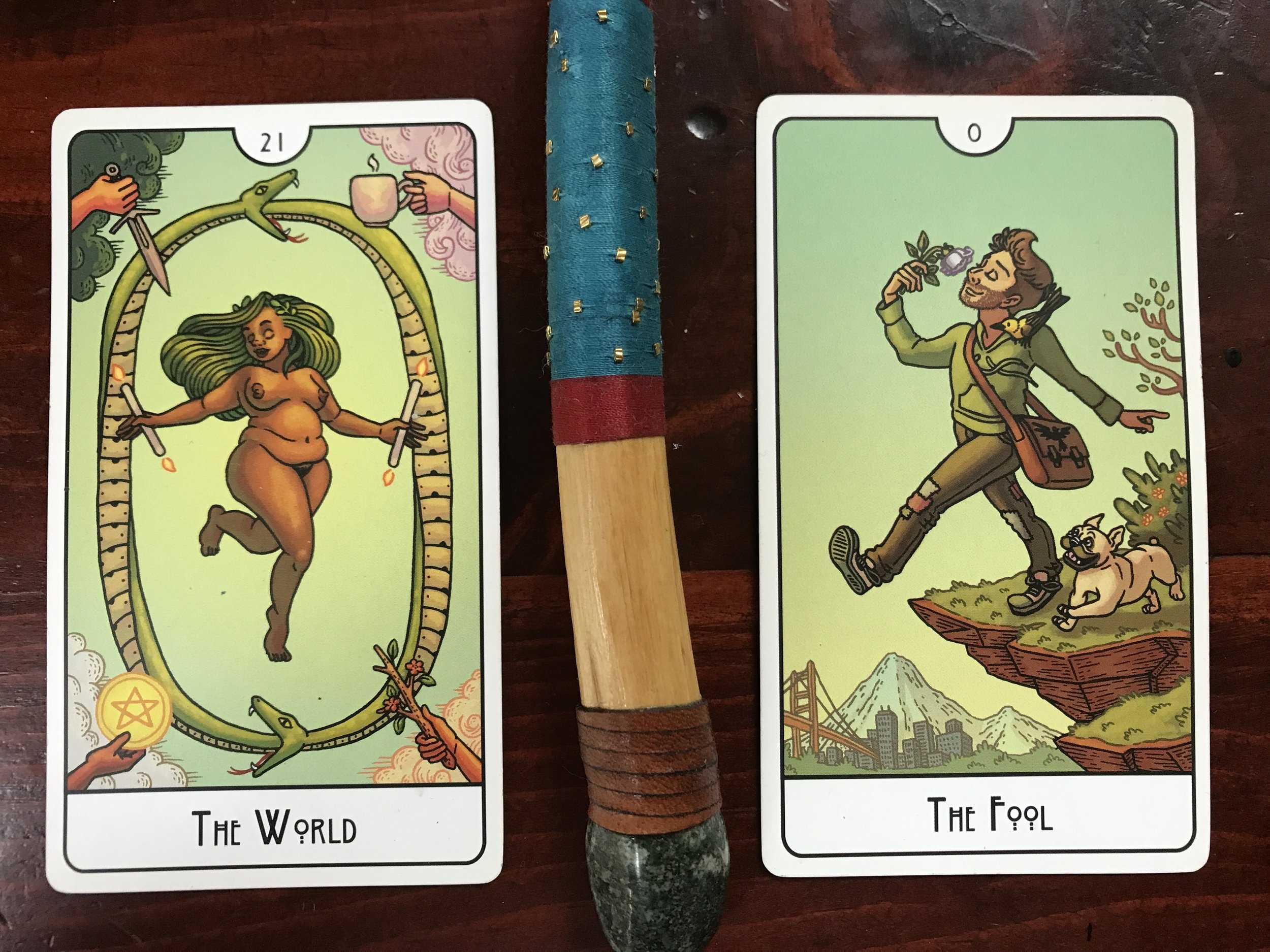 2 tarot cards separated by a pine wood wand tipped with a granite stone and wrapped in leather cord and red and blue thread against a dark wood background