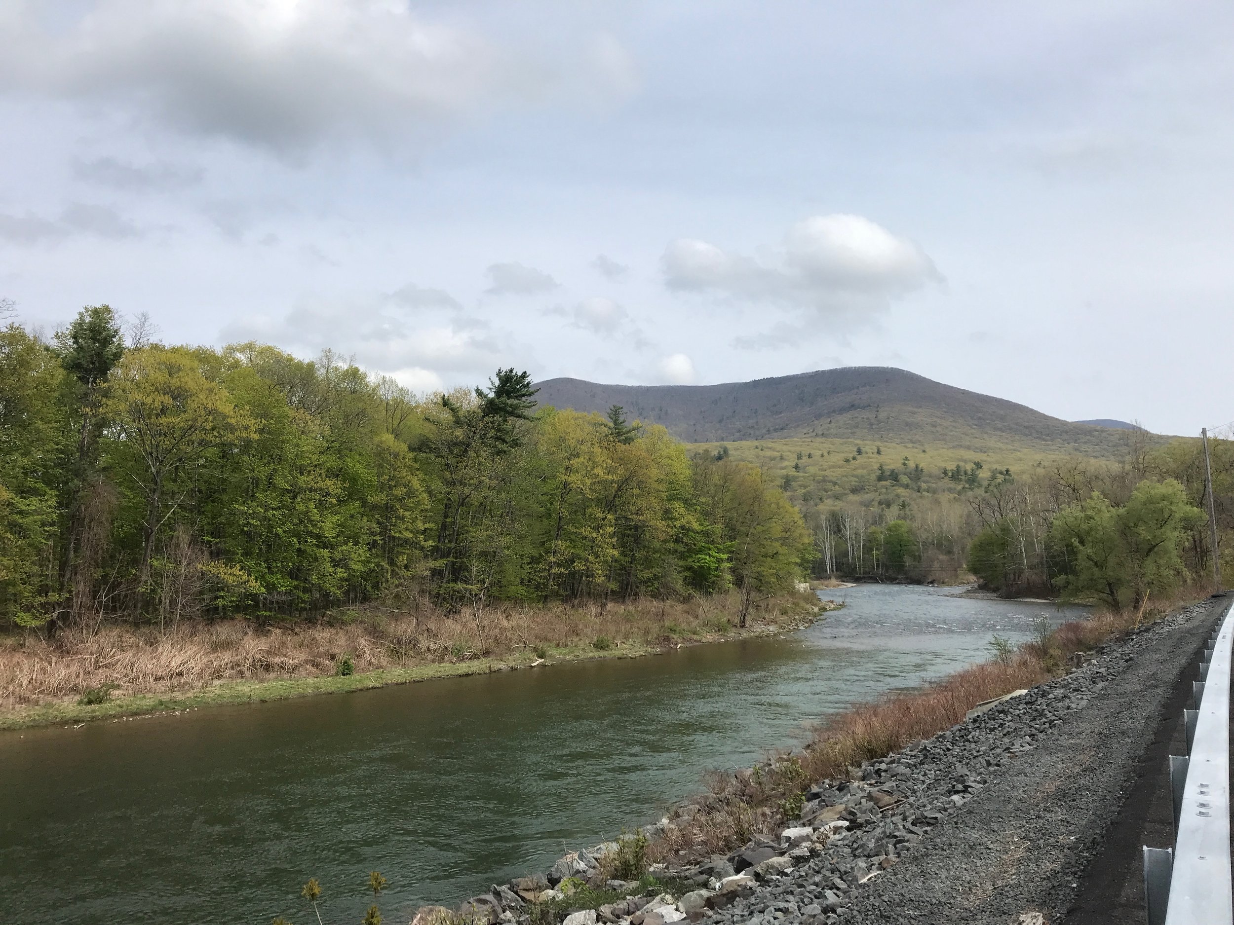 a river running along a road with a mountain in the distance up which the new green leaves of spring are creeping, the sky is pale blue with scattered clouds