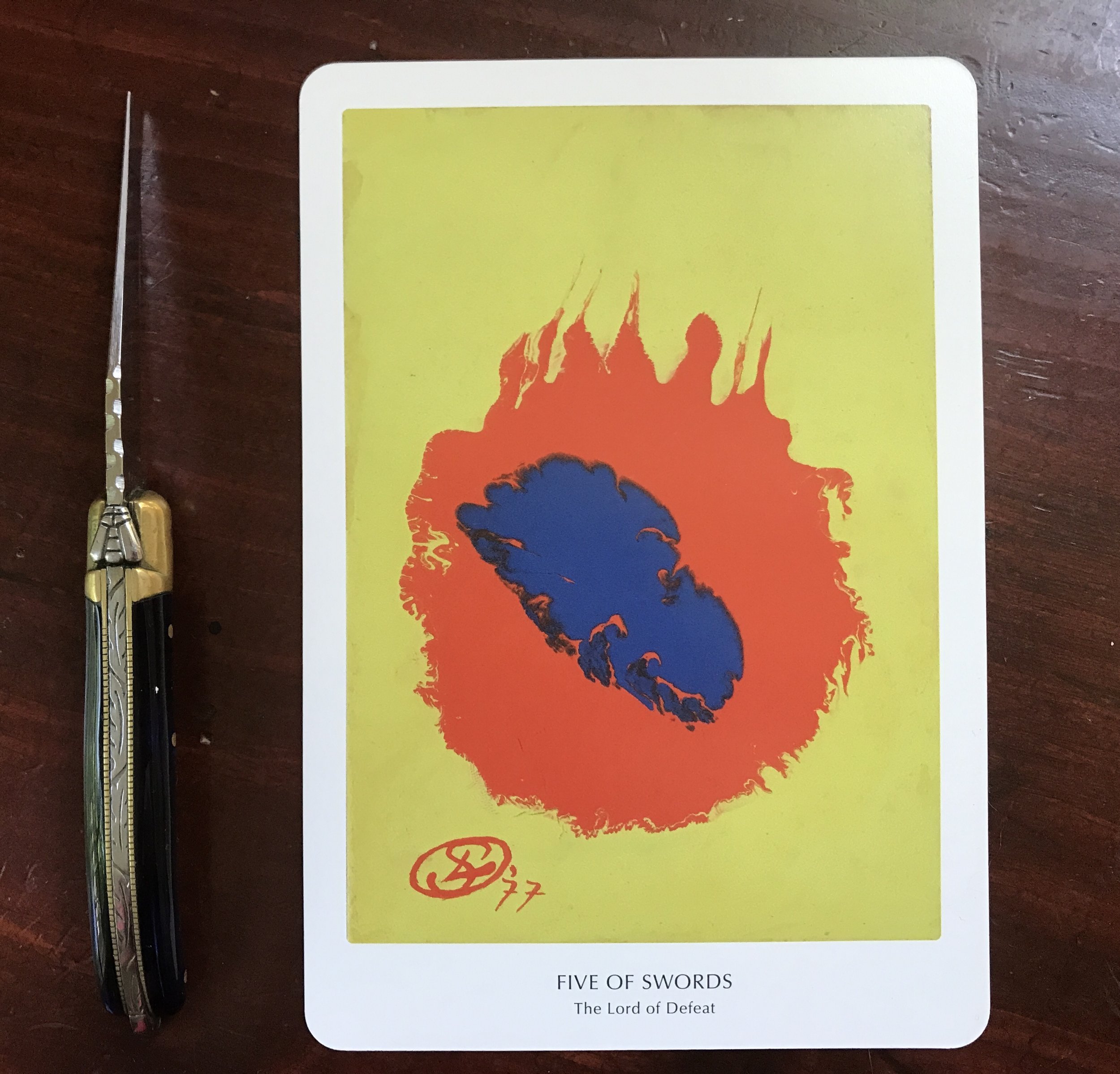 an abstract yellow, red and blue tarot card against a dark wood background with an extended pocket knife to the side on which there is a silver bee