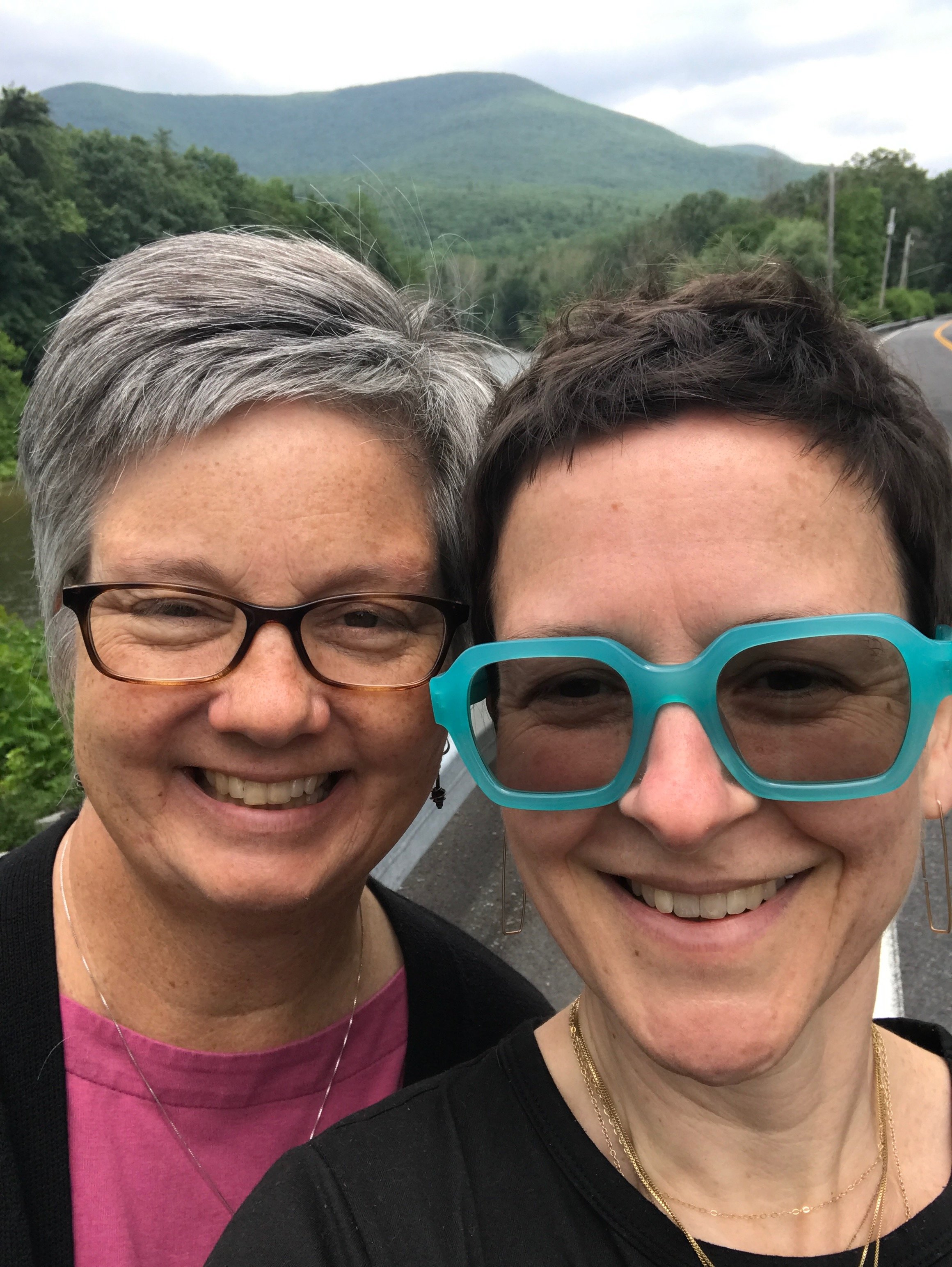 two white people, one with short grey hair and brown oval glasses wearing a pink shirt, one with short brown hair and square turquoise glasses wearing a black shirt