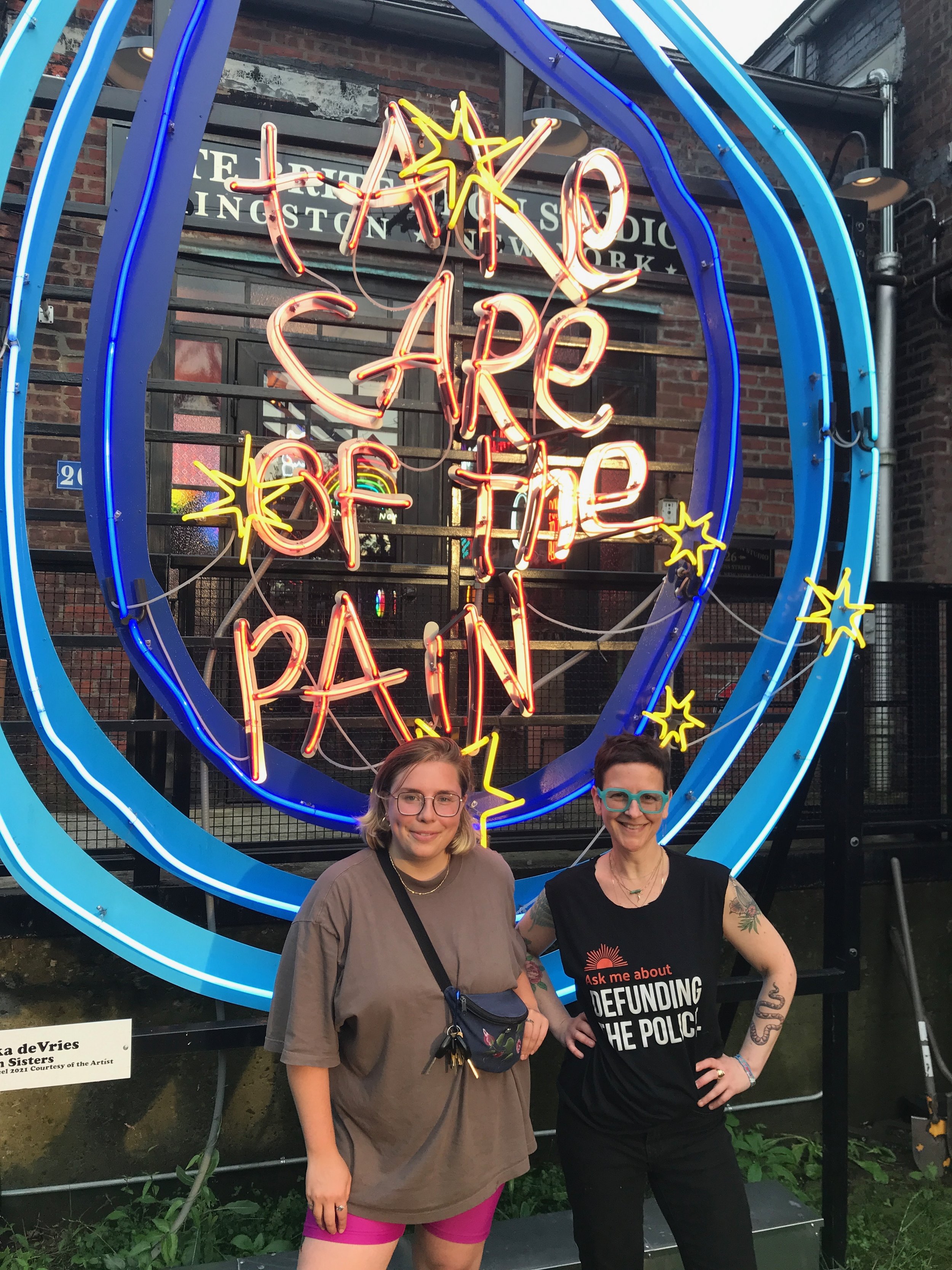 two figures stand in front of a neon artwork that says Take Care of the Pain