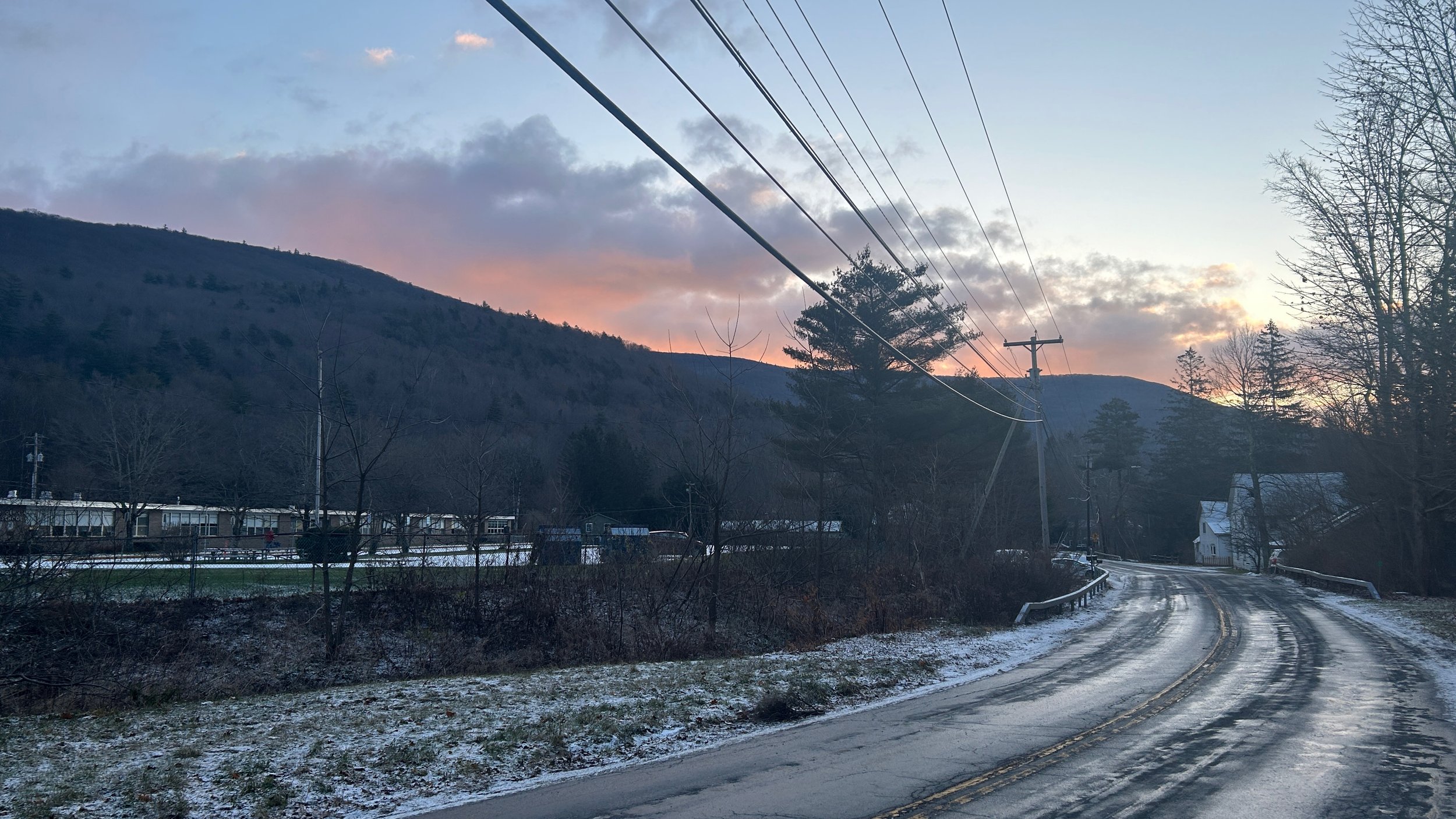 a rain-streaked two lane road with a snowy roadside and pink clouds over a winter-bare mountain