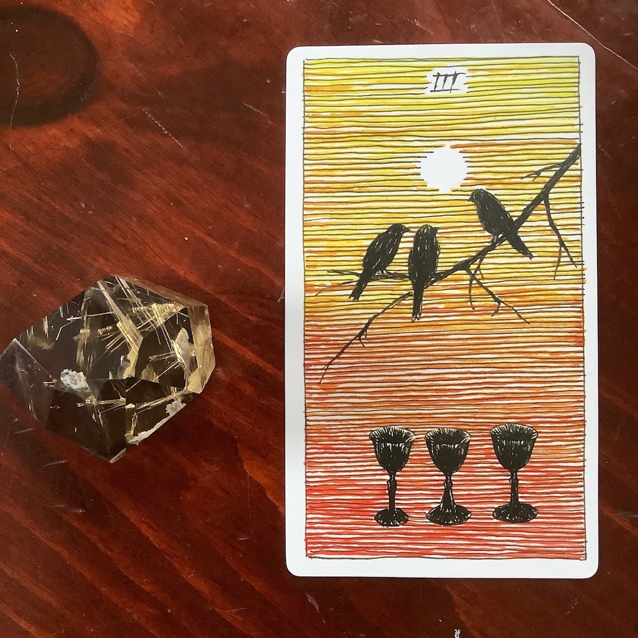 3 birds on a branch above 3 black goblets against an orange-to-yellow ombre ground with a white sun