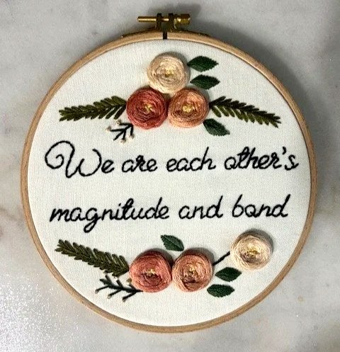 a circular hoop embroidery that says We are each other's magnitude and bond, with three pink flowers above and below
