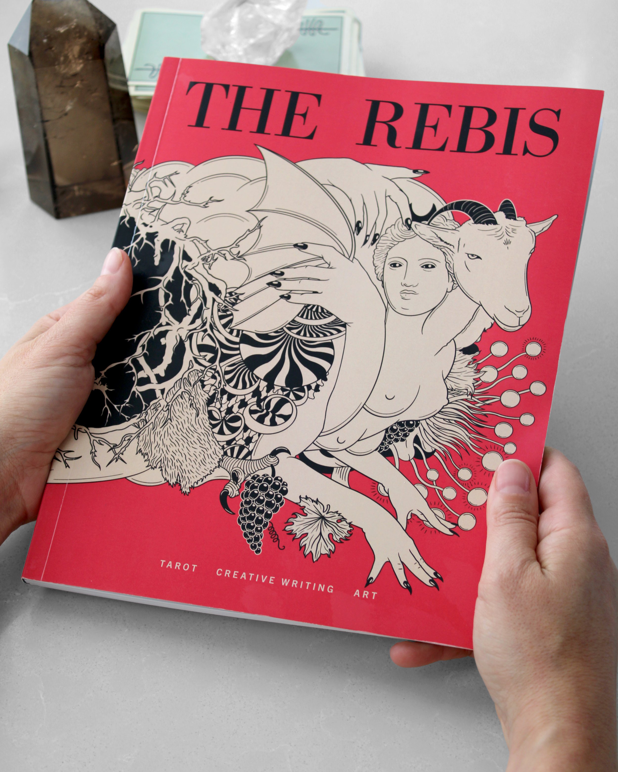 two hands hold a red journal that says The Rebis on top and tarot, creative writing and art on the bottom with an illustration showing a woman and goat creature