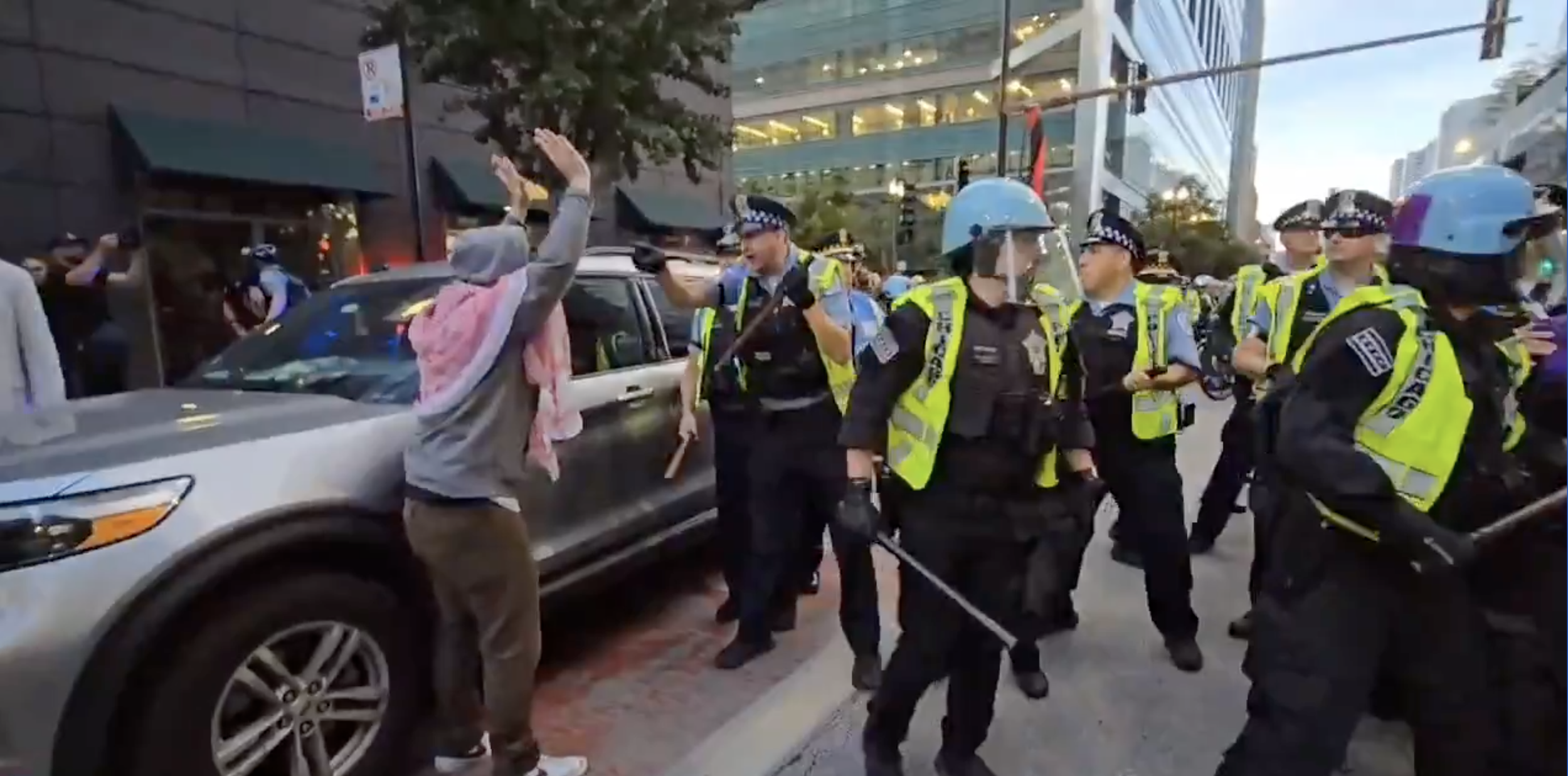 a protester standing in front of a car with both hands in the air and a crowd of neon yellow vested police officers with batons