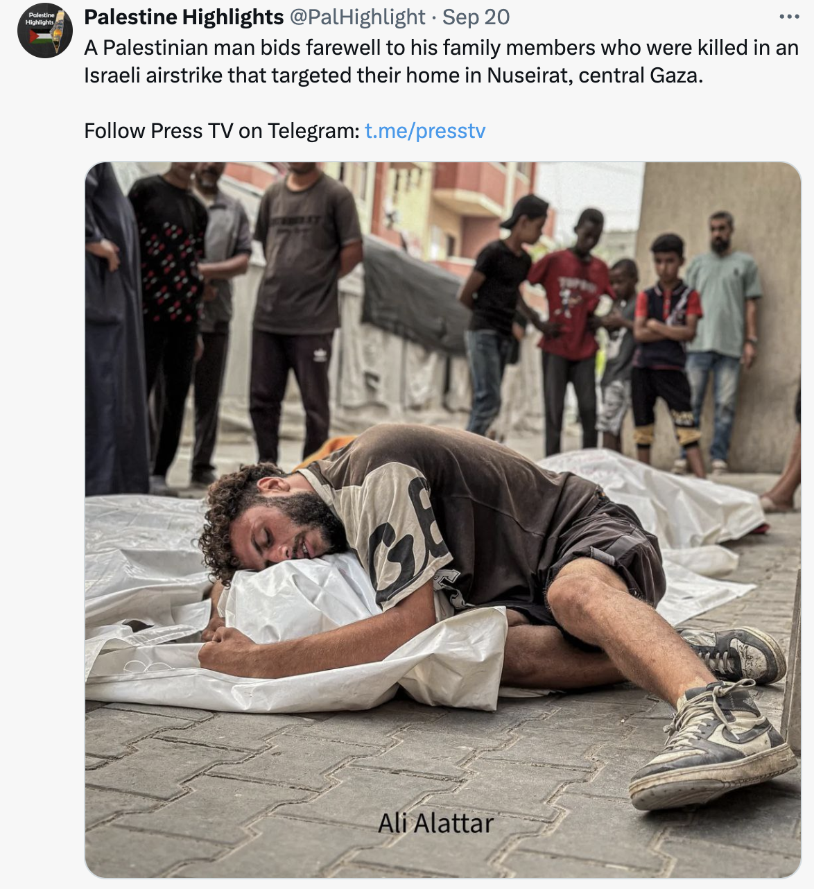 a bearded Palestinian man with curly hair wearing a t-shirt, shorts and dusty Nike high-tops cradles a body under a sheet of white plastic