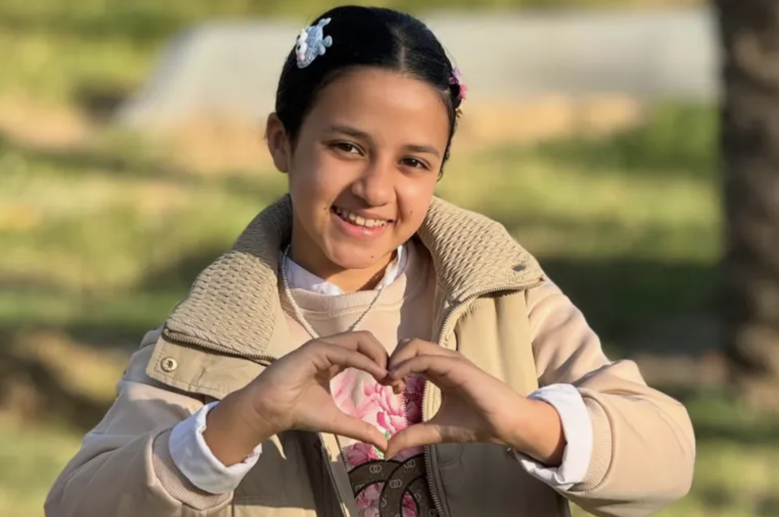 a smiling young Palestinian girl with a barrette in her parted black hair makes a heart gesture with her hands