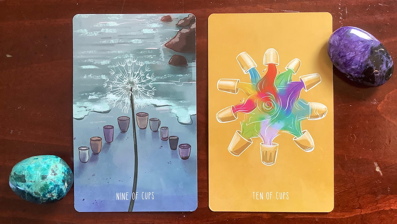 two colored stones flanking two tarot cards, one blue with 9 cups, the ocean's edge and a dandelion, the other gold with 10 gold cups pouring color into a swirling center