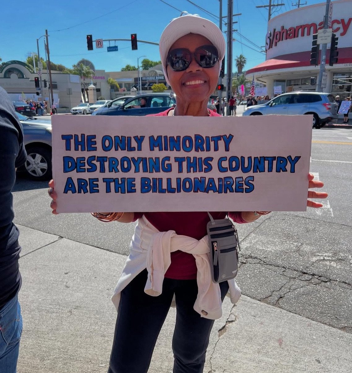 a thin black woman wearing a white hat and sunglasses smiles while holding a sign that says The only minority destroying this country are the billionaires