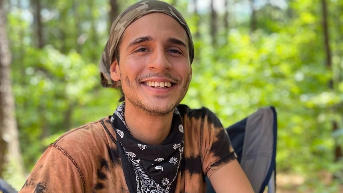 a racially ambiguous young person wearing a headwrap, tie-dyed t-shirt and dark bandana neck scarf smiles gorgeously with green trees in the background