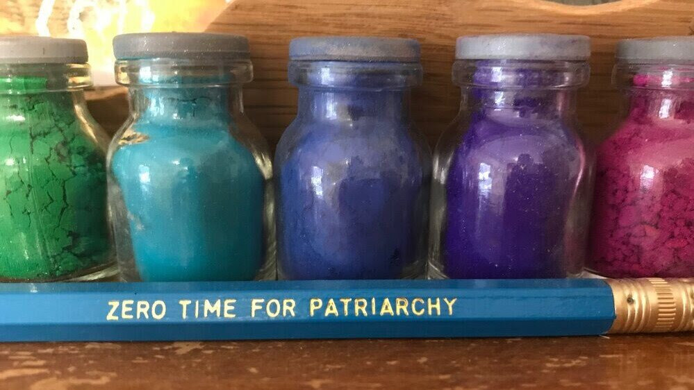 tiny glass jars of colored powdered pigment with a blue pencil that says in gold letters: zero time for patriarchy 