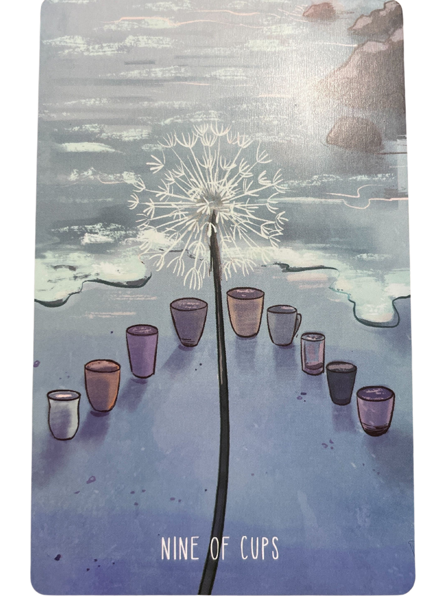 a tarot card showing a dandelion poof on a stem in front of an arrow-shaped arrangement of cups at the edge of the sea and sand