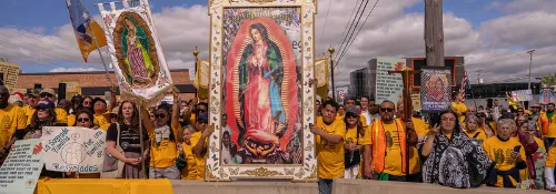 a crowd of protesters and clergy wearing bright yellow shirts holds up a large image of Our Lady of Guadalupe against a cloudy blue sky