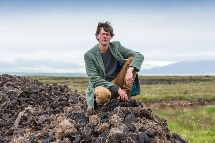 a white thin man faces the viewer wearing glasses, a green wool jacket, slate colored sweater and tan pants and boots crouches on a pile of peat with the Irish landscape in the background