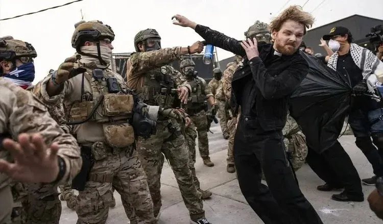 masked fascist jackboots wearing masks and military gear spray chemical weapons in the face of a white man with blonde hair and beard wearing all black and the collar of a clergyperson