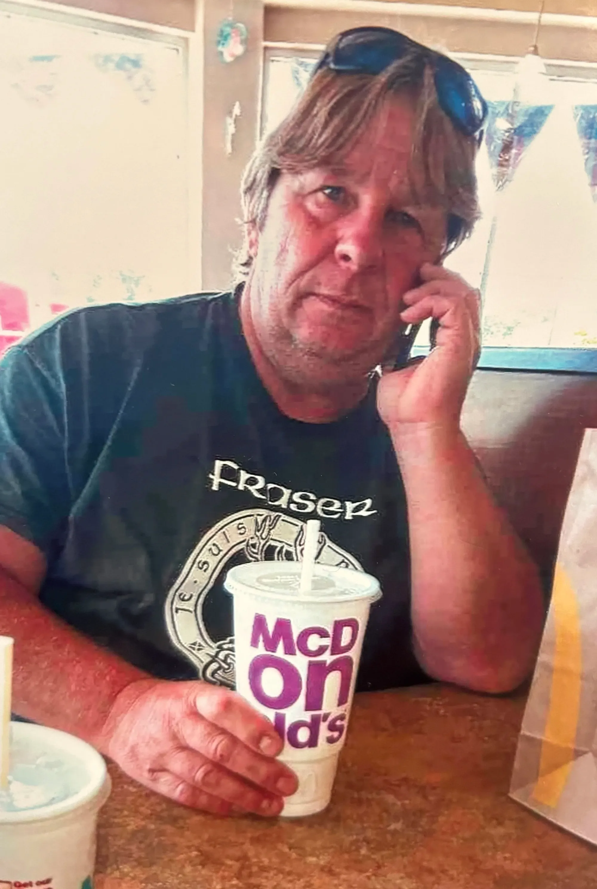 a big white guy in his late 50s with blonde hair and a cell phone pressed to his ear in one hand and a giant mcdonald's soft drink in the other