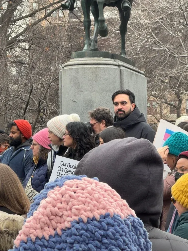 a bearded man in a black hooded winter coat standing amidst an outdoor crowd in front of a large monument