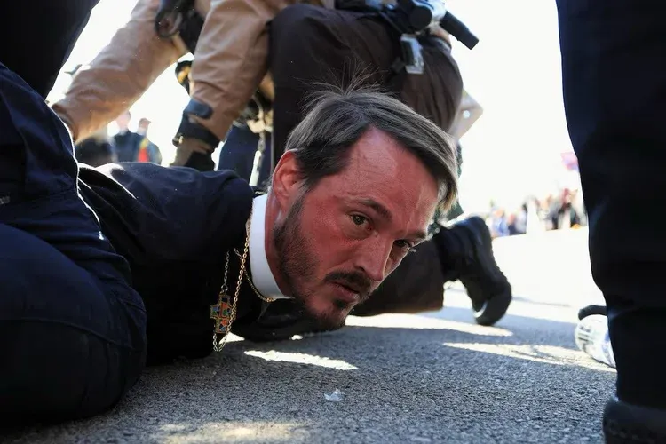 a white bearded man wearing a cross and clerical collar looking directly at the camera while being arrested on the ground by uniformed agents