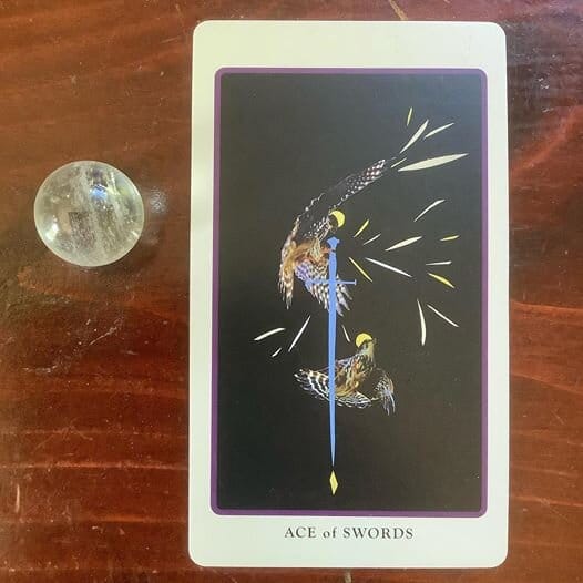 a quartz crystal sphere alongside a tarot card depicting a blue downward pointing sword and flying birds