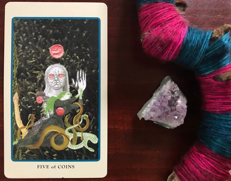 a tarot card showing a macabre figure, snakes and coins against a wood background next to which is an amethyst geode and a pink and blue string-wrapped stick