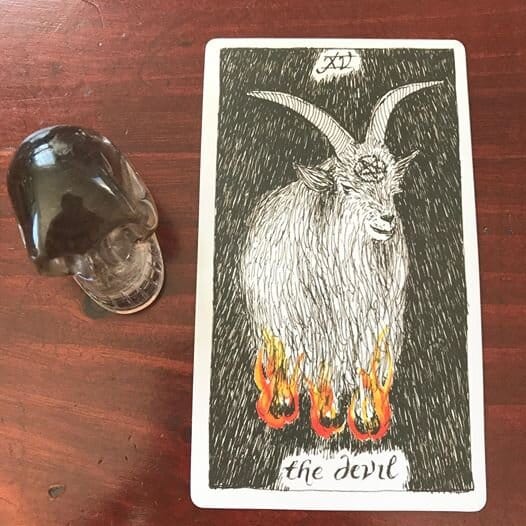 a line drawing of a long-horned goat with feet on fire and a pentacle on its forehead stands against a black background