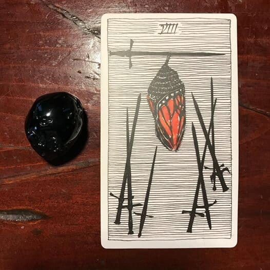 a tarot card of a monarch butterfly in a chrysalis hanging from a sword with 7 swords surrounding it against a ground of horizontal black lines
