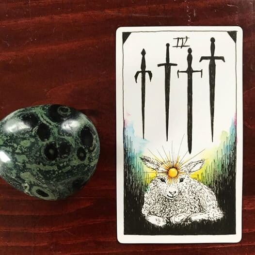 a lamb with a radiating sun at their third eye sits at the base of the card, 4 downward-pointing swords hover above