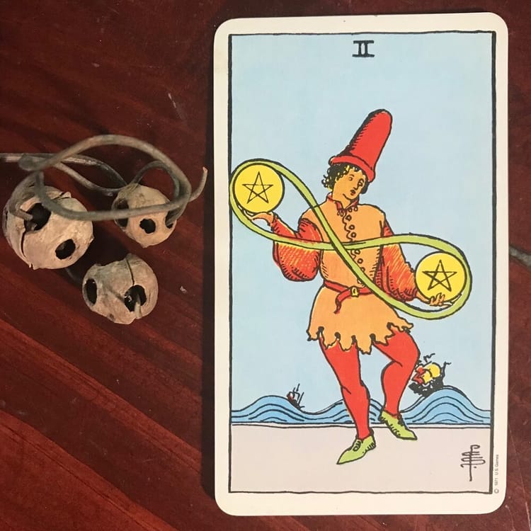 light brown seed pods through which a curly dried stem is threaded next to a tarot card of a figure in red and orange juggling 2 yellow pentacles