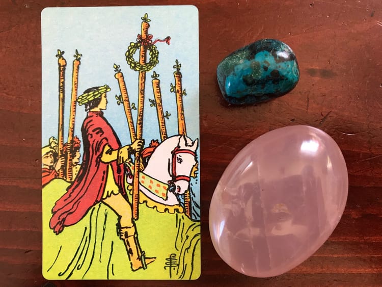 a tarot card of a figure wearing a laurel wreath crown riding a horse and two colored stones against a dark wood background