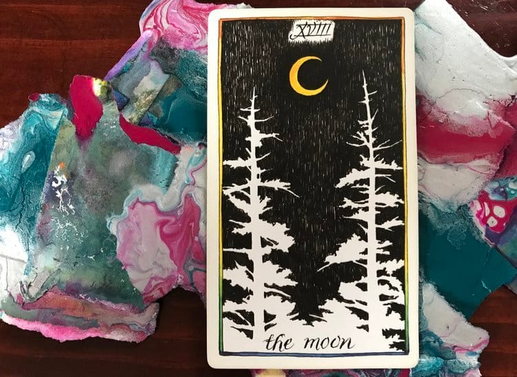 a black and white tarot card against a pile of abstract paintings against a dark wood surface